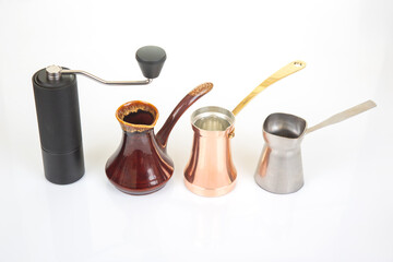 A black manual coffee grinder with a metallic handle stands beside a ceramic cezve, a copper cezve with a gold handle, and a stainless steel cezve on a white background