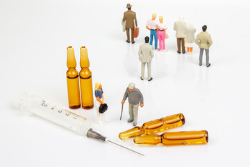 Miniature doctor and elderly figurine stand near three amber vials, a syringe, and a broken vial on a white surface, symbolizing healthcare, vaccination, or medical care concerns.