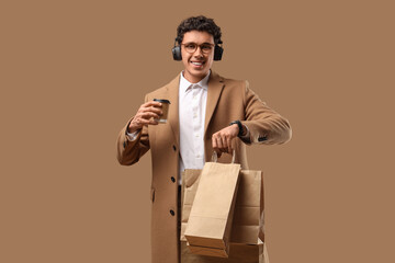 Stylish young man in headphones with coffee and shopping bags on beige background