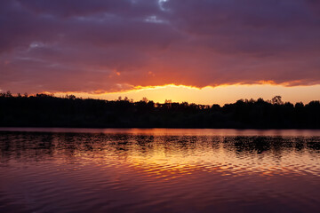 Serene lake at sunset with golden reflections on calm water, surrounded by lush trees under a dramatic sky. Perfect for nature and tranquility themes.
