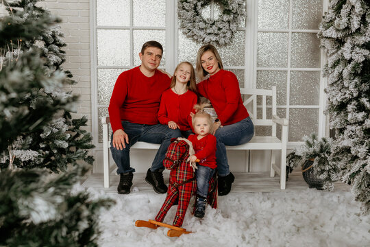 Family of four sitting on a white bench, with the youngest child riding a rocking horse, surrounded by snow and Christmas decorations - Powered by Adobe