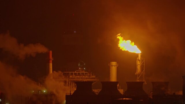 An industrial flare stack burns off excess gas with a large, bright orange flame and thick smoke against a dark night sky. Slow motion footage.