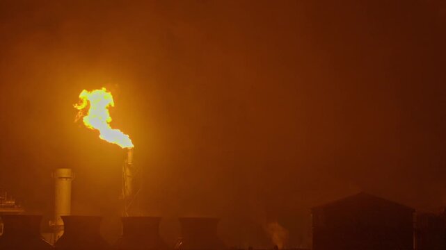 An industrial flare stack burns off excess gas with a large, bright orange flame and thick smoke against a dark night sky, highlighting energy production and potential environmental impact.