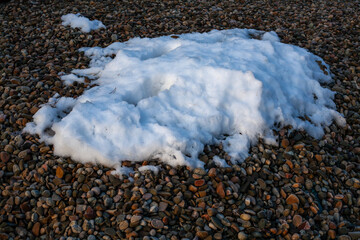 Patch of melting snow lying on a bed of smooth pebbles, creating a natural contrast of textures in winter light