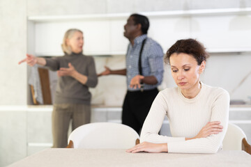 Adult sad woman sitting at table during family quarrel between elderly woman and adult man..