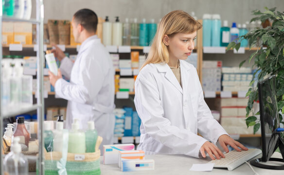 Female pharmacist works at a computer, introduces the presence of new packages with medicines against the background of a male pharmacist. European pharmacy workers near the counter