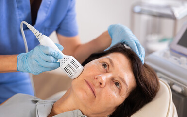 Aged woman undergoing hardware facial treatment using cold hammer helping to shrink pores, repair damaged skin and relieve inflammation in modern aesthetic medicine clinic
