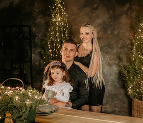 Happy stylish family of three sitting at table during Christmas dinner with festive decor and lights in the background