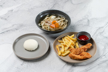 A vibrant Asian fusion meal showcases a bowl of noodles with vegetables, crispy fried chicken with fries, and a soft steamed bun. The setting highlights modern dining aesthetics