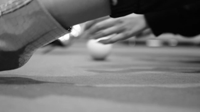 Close-up black and white shot of hand spinning cue ball on felt, racking focus to blurred woman wearing shiny silver high heel boots in background.

