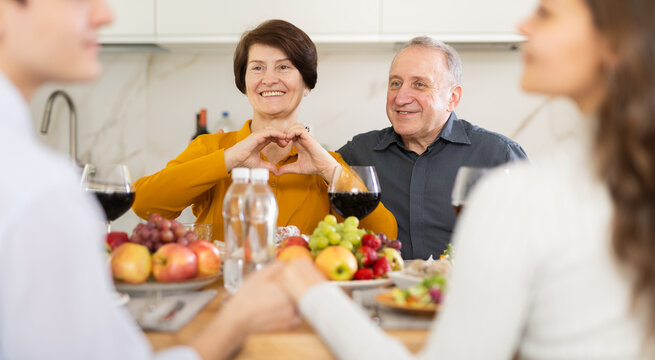 Senior parents showing heart gesture, happy with love of adult son, sitting at set table with girlfriend during cozy family dinner at home - Powered by Adobe