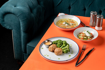 A colorful dining experience awaits on a bright orange table. Tasty soup brimming with herbs, fresh salad, and juicy meatballs are served alongside broccoli, creating an inviting atmosphere