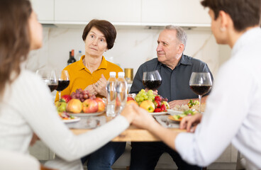 Puzzled elderly mother looking on with concern and surprise while adult son in love engaging in conversation with girlfriend at dinner table during family meal at home, young couple in love clasping