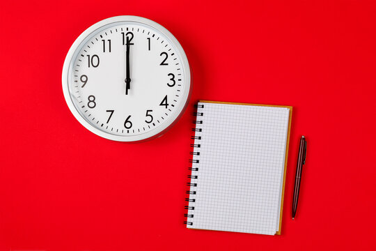 Round clock pointing to 12:00 and an empty notebook. New Year is time for new decisions, countdown, and planning. Planning and deadline concept on a red background.