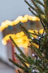 Close-up of a Christmas tree with string lights, with warm festive bokeh from a carousel or holiday lights in the blurred background