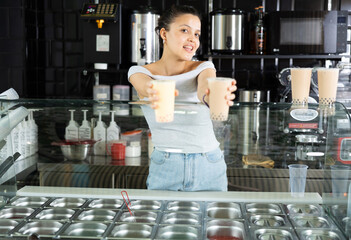 Happy young Asian woman behind glass counter holding out two bubble tea drinks in modern beverage shop, showcasing friendly service..