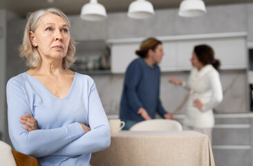Frustrated senior woman is sitting in kitchen during quarrel between her son and his wife. Complicated family relationships, daughter-in-law and husband yell at each other