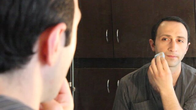 A man sits at a dressing table, looking in the mirror while applying makeup with a brush to his eyebrows. He then checks his phone briefly.


