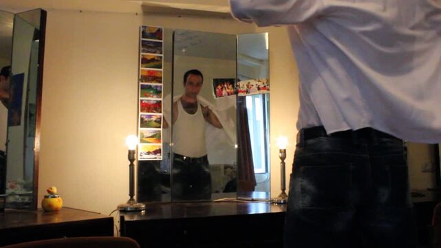 A man stands before a dressing table mirror wearing an undershirt and jeans. He puts on a white dress shirt, getting ready for work or an event.

