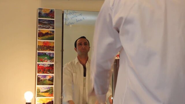 A man stands before a dressing table mirror wearing an undershirt and jeans. He puts on a white dress shirt, getting ready for work or an event.

