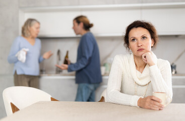 Woman is sitting in kitchen and is upset by quarrel between her boyfriend and elderly mother-in-law. Mature mother-in-law screams and insults middle-aged male.