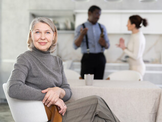 Obraz premium Glad mature woman sitting in the kitchen and middle-aged man and woman arguing angrily standing behind
