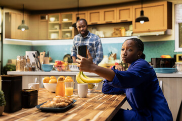 African american woman takes selfies on her smartphone and acting silly, boyfriend laughing at her and enjoying a cute funny moment together on a weekend morning. Relaxation at home.