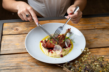 A person sits at a rustic wooden table, delicately cutting into a beautifully plated chicken dish. Garnished with pomegranate seeds and radish, this meal promises an explosion of flavor