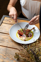 A person cuts into a beautifully presented dish of duck confit, served on polenta with colorful garnishes. The cozy ambiance enhances the dining experience