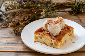 A delightful slice of warm apple cake topped with three scoops of creamy ice cream and crushed nuts, placed on a rustic wooden table with dried flowers in the background
