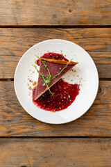 A carefully plated dessert showcases a slice of rich cake topped with a bright raspberry sauce. Fresh herbs add a touch of elegance to the presentation on a weathered wooden table