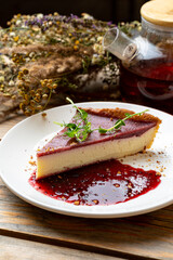 A luscious slice of cheesecake sits on a rustic wooden table, drizzled with vibrant berry sauce. A pot of herbal tea rests beside it, surrounded by dried flowers, creating a cozy atmosphere