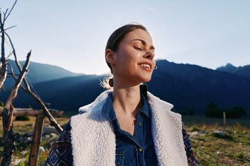 Fototapeta premium Woman portrait in mountains nature at golden sunlight, smiling and happy outdoors with casual jacket and wool vest, scenic landscape background and peaceful rural vibe during sunset.
