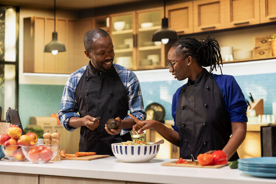 Young black man cutting an avocado to add within fresh salad, his girlfriend chopping other vegetables and mixing ingredients in a bowl. Partners with aprons preparing vegetarian lunch. - Powered by Adobe