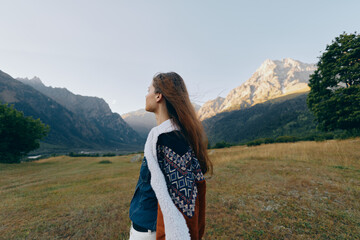 Fototapeta premium Woman in a meadow looks toward mountains in a wide landscape, wearing a patterned shawl; travel portrait of nature, solitude and serene outdoor exploration in golden light.