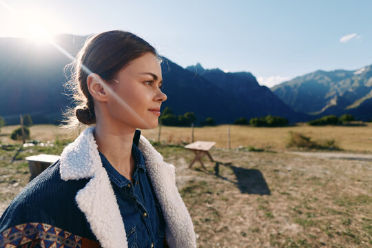 Woman portrait profile outdoors in mountain field with sunlight lens flare. Young female in shearling jacket looking contemplative at nature landscape with clear sky and peaks.