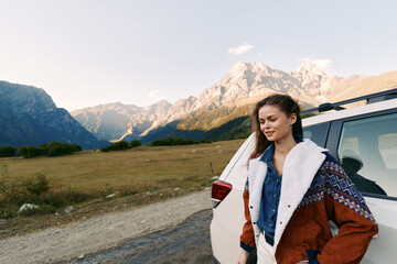 Fototapeta premium Woman by car on mountain road during outdoor travel, scenic landscape and nature view. Young female in warm jacket stands beside vehicle with peaks in background and calm expression