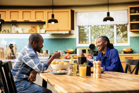 African american lovers clinking mugs with coffee in the morning, enjoying weekend bonding and sitting together. Sweet affectionate man and woman chatting at the breakfast table. - Powered by Adobe