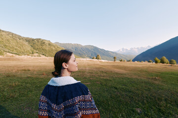 Fototapeta premium Woman stands in meadow valley looking at mountains and nature landscape outdoors, travel portrait in sweater with serene scenery, grass, sky and panoramic alpine view.