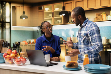 Joyful African American couple preparing breakfast together with coffee cups in bright kitchen. Checking a homemade pastry recipe for a healthy organic meal in the morning. Warmth at home.