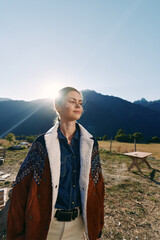 Fototapeta premium Woman portrait outdoors in mountains with sunlight at sunrise, wearing denim shirt and shearling jacket in countryside. Serene profile in nature meadow, casual fashion and confident travel mood.