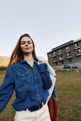 Fototapeta premium Woman smiling in denim shirt holding a jacket outdoors in a grassy field near a residential building, casual portrait with relaxed pose, natural light and confident expression.