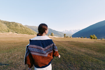 Fototapeta premium Woman in a cozy sweater walking across a mountain meadow, nature landscape with scenic outdoors scenery. Back view portrait captures calm hiking journey and tranquil horizon.