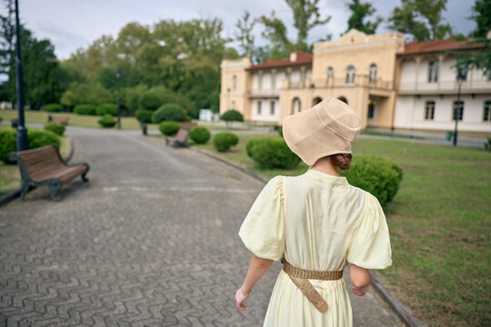 Elegant woman in a light dress and woven hat walks along a courtyard path toward a historic estate, creating a serene vintage ambiance and timeless travel atmosphere with soft sunlight - Powered by Adobe
