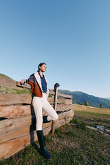 Fototapeta premium Woman leaning on wooden fence in mountains countryside meadow under clear blue sky, wearing jacket and boots, full body portrait in rural outdoors setting for fashion and travel.