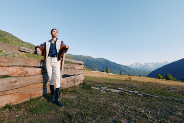 Fototapeta premium Woman leans on wooden fence in alpine meadow against distant mountains, countryside travel scene. Casual outfit with boots, relaxed portrait in wide open landscape under clear blue sky.
