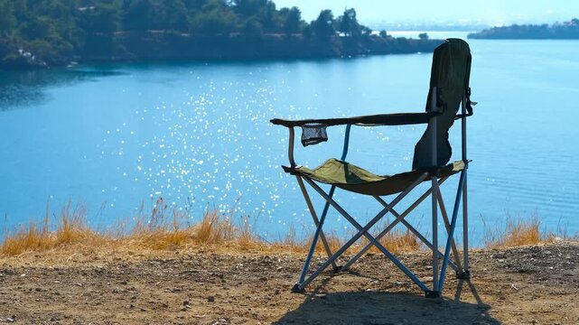 Peace of mind on a camping holiday. Empty camping chair rests on a hill, providing a stunning view of a sparkling blue lake surrounded by lush trees. The scene evokes tranquility and relaxation
