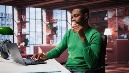African american adult typing on a computer in his loft apartment, engaging in online freelancing work and multitasking efficiently. Flexibility of remote work in a cozy home office. Camera B.