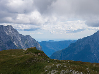 A panoramic alpine landscape of Montgart in the Alps featuring rugged rocky cliffs, soft green slopes, a winding mountain road and a dramatic cloudy sky