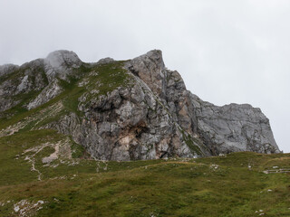 A panoramic alpine landscape of Montgart in the Alps featuring rugged rocky cliffs, soft green slopes, a winding mountain road and a dramatic cloudy sky